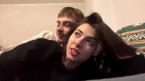 Media: Video of a young couple lying on a bed, the woman with long dark hair and a black top, the man with short light hair, both looking relaxed.
