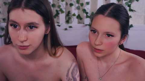 Video of two topless women with pale skin and dark hair, one with a tattoo, sitting close, indoors with green vines and white walls in the background.