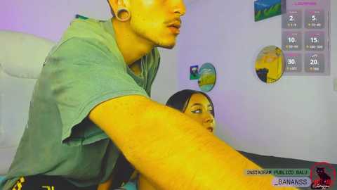 A CGI screenshot from \"Instagram Live: Banana Rass,\" featuring a man with tanned skin and a woman with fair skin, both wearing casual clothes, in a modern room with a digital clock and colorful wall art.