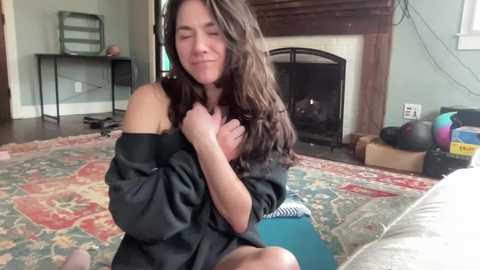 Video of a young woman with long brown hair, wearing a black off-shoulder top, sitting on a colorful rug, smiling, in a cozy living room with a fireplace.
