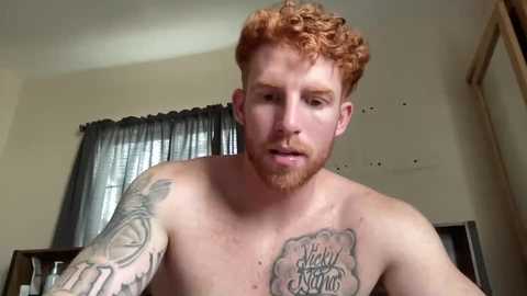 Video of a shirtless, muscular, red-haired man with a beard, tattoos, and a pierced ear, indoors near a window with a curtain.