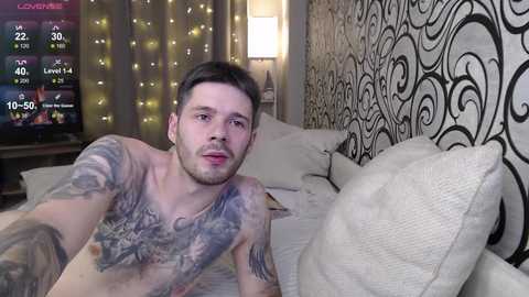 Video of a fit, tattooed, young man with short brown hair, lying shirtless on a bed in a modern bedroom with black and white patterned wallpaper and a TV screen showing a video game.
