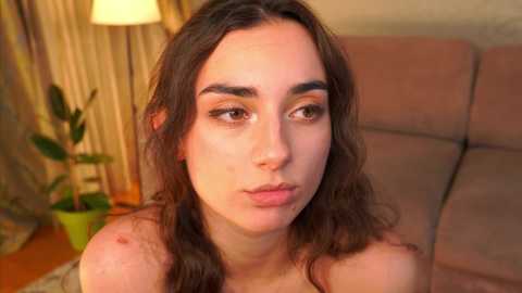 Video of a young woman with fair skin and long brown hair, puckering her lips, in a cozy living room with beige curtains, a green plant, and a lit lamp.