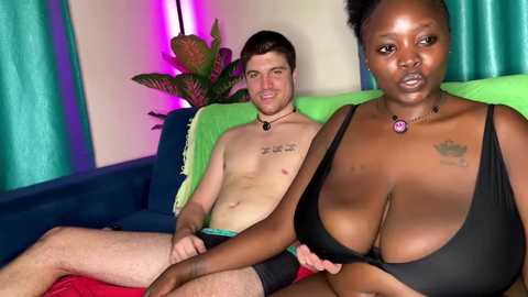 Video of a light-skinned, shirtless man with short brown hair, wearing black underwear, sitting on a couch with a dark-skinned woman with large breasts, wearing a black bra, in a room with green curtains and a potted plant.