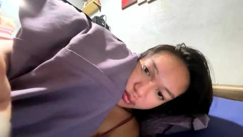 Video of a young East Asian woman with medium-length black hair, wearing a light purple hospital gown, lying on a blue hospital bed, looking tired, in a dimly lit room with white walls and medical equipment.