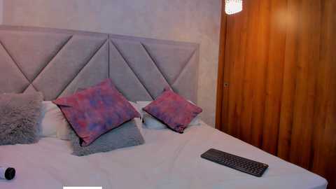 Video of a modern bedroom with a light grey, upholstered, geometric-patterned headboard, two purple and pink patterned pillows, a white bedsheet, a black keyboard on the bed, and a wooden door.