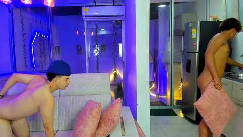 Video of two young Asian men, one nude and wearing a blue cap, the other shirtless, standing in a modern apartment with a large refrigerator and glass door, under purple and blue lighting.