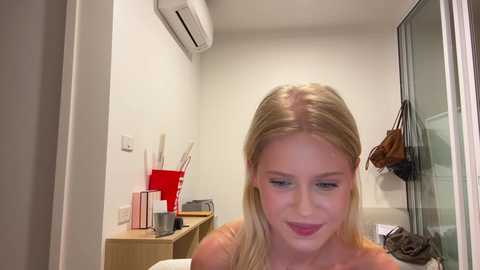 Video of a blonde woman with fair skin, wearing a red top, smiling, in a modern, minimalistic bedroom with white walls, an air conditioner, and a wooden cabinet.