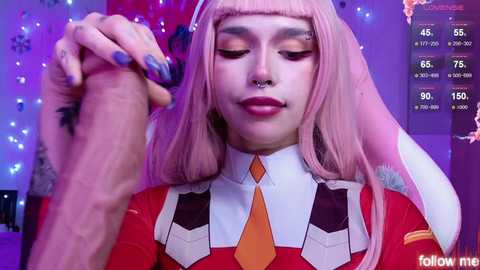 Video of a young woman with pale skin, long pink hair, and bangs, wearing a red and white futuristic costume with an orange tie, holding a hand. Background features blue and purple lighting.