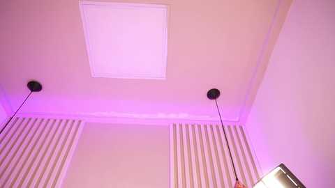 A video of a corner room with beige walls, a rectangular ceiling light, and two black wall-mounted speakers emitting purple light. Vertical white-striped curtains are partially visible.