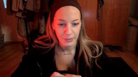 Video of a fair-skinned woman with long brown hair, wearing a black headband and top, looking at her phone. Background shows wooden wardrobe, hanging clothes, and a red glow.