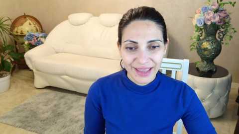 Video of a smiling woman with dark hair in a blue shirt, seated in a cozy living room with a white couch, floral arrangement, and potted plants.