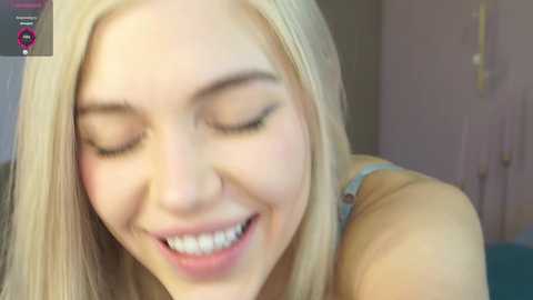 Video of a smiling, fair-skinned, blonde woman with closed eyes, wearing a light blue top, in a softly lit room with muted pastel walls.