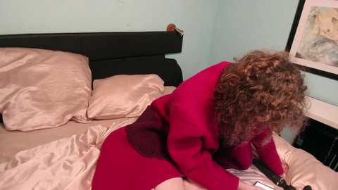 Video of a woman with curly brown hair, wearing a red dress, lying on a beige bed with a black headboard. She's reading a book, surrounded by soft lighting and framed artwork on the pale blue wall.