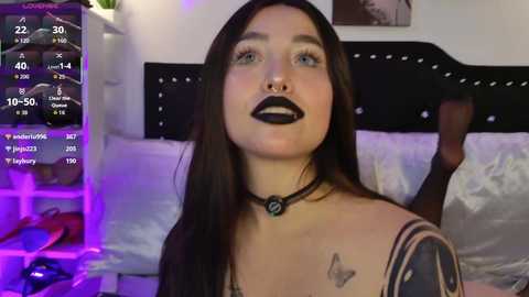 Video of a woman with long dark hair, black lipstick, and tattoos, wearing a black choker, in a dimly lit bedroom with purple lighting.
