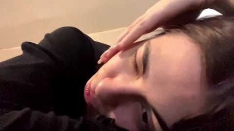 Video of a young woman with fair skin, resting her head on her hand, eyes closed, lying on a black blanket. She wears a black sweater. The background is a beige wall.