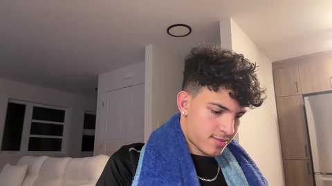 Media: Video of a young man with curly dark hair, wearing a black shirt and a blue towel, standing in a modern, dimly lit living room with white furniture and a refrigerator.