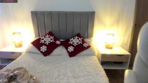 Video of a modern, cozy bedroom with a gray upholstered headboard, a white bedspread, and two red pillowcases adorned with white snowflakes. Nightstands with warm-toned lamps flank the bed. The room's decor is minimalistic and inviting.