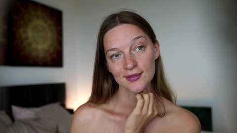 Video of a fair-skinned, light-haired woman with freckles, smiling, with a relaxed expression. She has blue eyes and is topless, posing in a softly lit bedroom with a bed and wall art in the background.