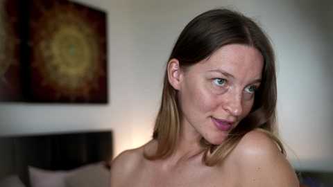 Video of a fair-skinned woman with straight, shoulder-length brown hair, green eyes, and a subtle smile. She is topless, set against a modern, dimly-lit bedroom with abstract art on the wall.