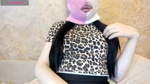 A video of a woman with pale skin, wearing a leopard print top, black pants, and a pink knit hat with a face cutout. She sits against a textured beige wall, her mouth open.