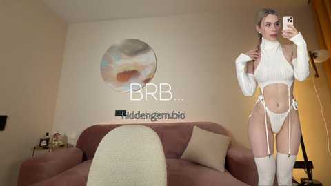 Video of a blonde woman with a slim physique, wearing a white, high-necked crop top and matching high-waisted panties, posing in a modern bedroom with a circular abstract wall art and a beige couch.