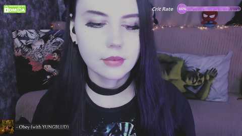 A video of a pale-skinned woman with long black hair, wearing a black choker and a Batman shirt, sitting in a dimly lit room with Spider-Man and Batman posters on the wall.