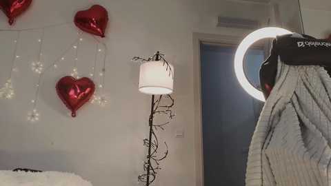Video of a modern bedroom with a white wall, red heart-shaped balloons, a white lamp, and a gray sweater hanging on a black hanger.
