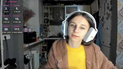 Video of a young woman in a cozy, cluttered kitchen wearing large white headphones, a yellow shirt, and a brown fleece jacket.