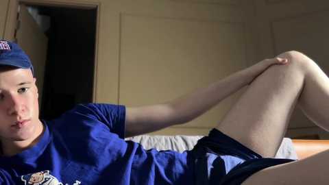 Video of a young man with fair skin, wearing a blue t-shirt and cap, lying on a bed, stretching his leg, in a dimly lit, beige-walled room.