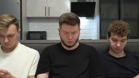 Video of three young men sitting closely on a gray couch in a modern kitchen with white cabinets and black appliances. They wear casual black and white t-shirts, focused on their phones.