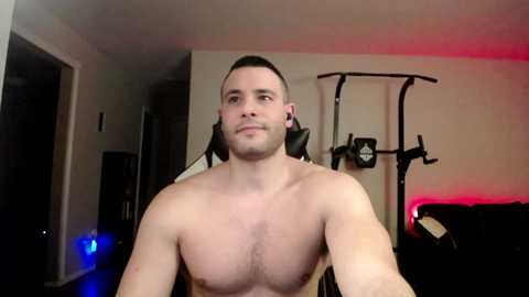 Video of a shirtless, muscular, light-skinned man with short dark hair, sitting on a black exercise machine in a dimly lit room.