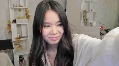 Video of a young Asian woman with long, dark hair, wearing a white robe, smiling while posing in a modern, minimalistic bathroom with white walls, shelves, and plants.