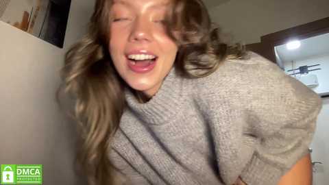 Video of a smiling woman with wavy, light brown hair wearing a gray knitted sweater, indoors with beige walls and a ceiling fan visible in the background.