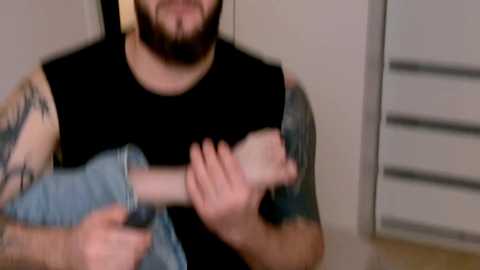 Media: Video of a bearded man with tattoos on his arms, wearing a black sleeveless shirt, holding a limp, bloodied arm, in a dimly lit room with a white dresser.