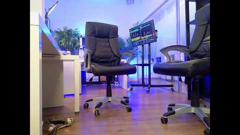 Video of a modern broadcast studio with two black leather chairs, a desk, and a large monitor displaying colorful graphs, under blue lighting.