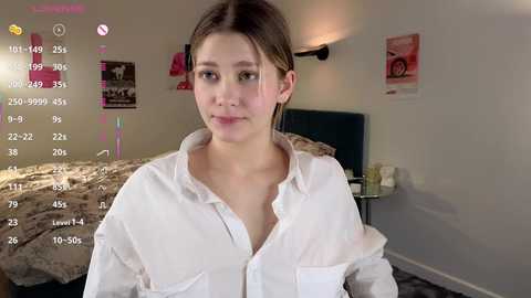 Media: A young, light-skinned woman with brown hair, wearing a white, unbuttoned shirt, sits in a modern, minimalist bedroom. The background includes a bed with a beige duvet, posters, and a lamp. The image appears to be a selfie with a virtual camera overlay.