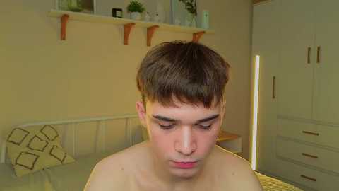 Video of a young, shirtless Caucasian boy with short, dark hair, looking down, in a softly lit, minimalistic bedroom with beige walls and wooden shelving.
