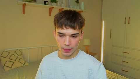 A video of a young man with short brown hair, wearing a white shirt, standing in a bedroom with beige walls, a bed, and a white wardrobe in the background.
