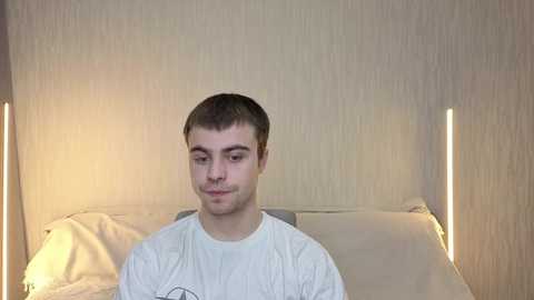 Video of a young man with short brown hair and a light beard, wearing a white T-shirt, sitting on a bed with beige pillows and a white blanket. The background has a beige textured wall and two tall, narrow lamps.