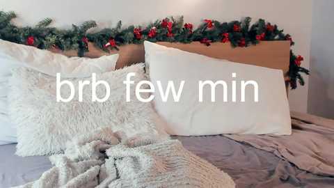 Video of a cozy bed with a white pillow, beige blanket, and a soft, textured gray throw, adorned with a festive garland of greenery and red berries. Text overlay reads \"brb few min.\