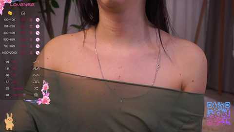 A close-up video of a fair-skinned woman with dark hair, wearing a sheer green top revealing her shoulders and chest. The background includes a potted plant and a blurred room.