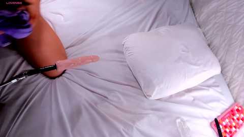 Video of a person with medium brown skin and purple hair, lying on a white bed with a pink vibrator between their legs, and a pink checkered box nearby.