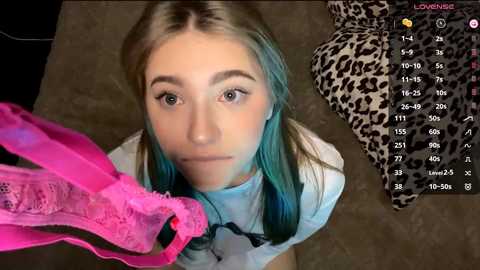 A video of a blonde woman with blue hair, wearing a white top, holding a pink lace bra, against a leopard-print background, with a live-streaming overlay showing viewers' chat messages.