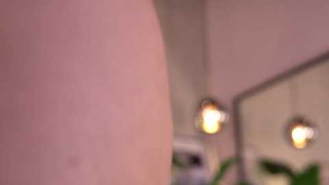 A blurred video of a person's cheek with a pale complexion, reflected in a mirror with two lit, round pendant lights in the background.