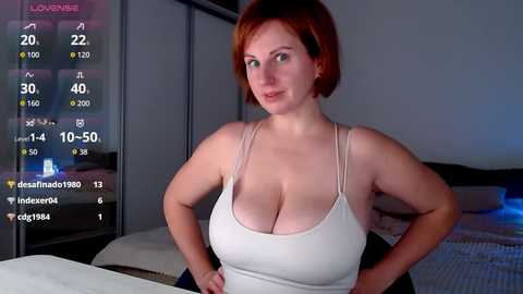 Video of a fair-skinned woman with red hair, wearing a low-cut white tank top, standing confidently in a bedroom. Background shows a bed and digital temperature and humidity readings.
