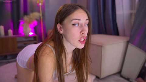 Video of a slender, young woman with long brown hair, wearing a white crop top and shorts, posing provocatively on a beige couch in a dimly lit room with purple and pink lighting.