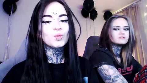 Video of two tattooed women with dark hair, wearing black shirts, puckering their lips, in a room with black balloons, gray curtains, and a gaming chair.