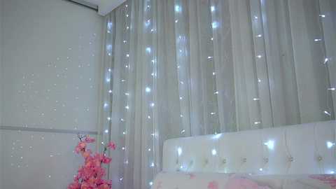 A video of a softly lit bedroom with white curtains adorned with twinkling fairy lights, pink flowers in a vase on a white bed, and a serene, dreamy atmosphere.