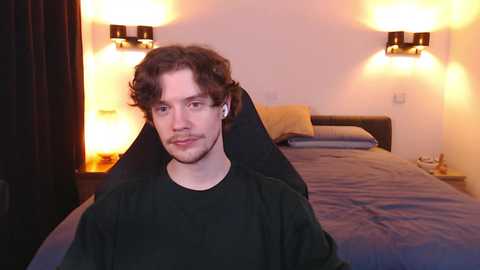 Video of a young man with curly brown hair, fair skin, and a light beard, wearing a black t-shirt, sitting in a dimly-lit bedroom with a neatly made bed, two wall-mounted lamps, and dark curtains.